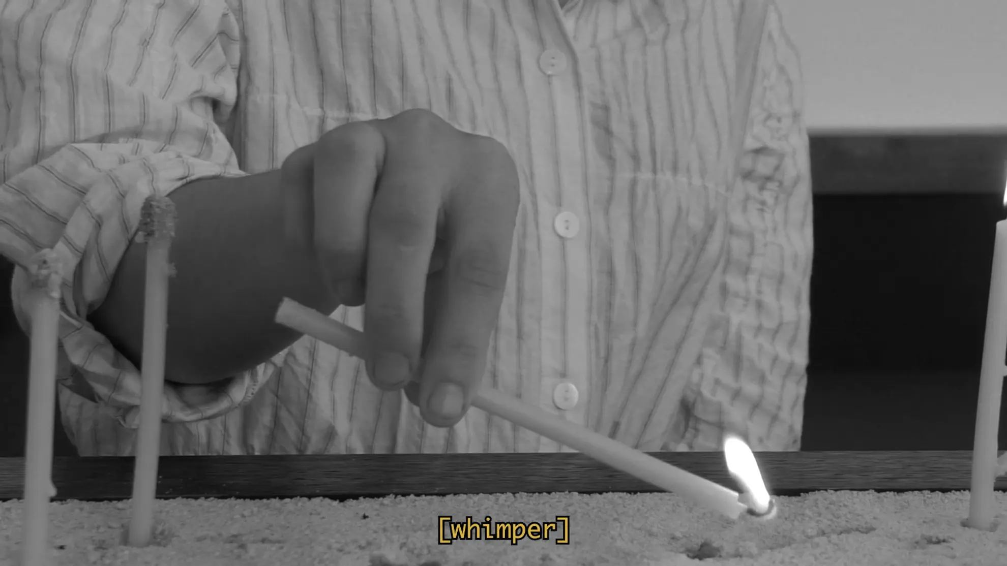 Close-up of a hand holding a thin lit candle pointing the flame downwards into a pile of sand. A caption in yellow text reads the word whimper in square brackets.