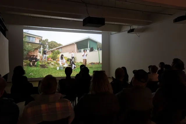 A dark room hosts a crowd of people watching the projector screen. The projector displays six people in a large grassy backyard of a red brick house, the people are all engaging in various activities.