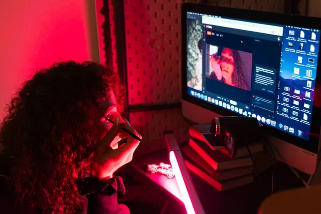 A woman sitting in front of a computer bathed in red light uses a webcam reflection to do her eyeliner.