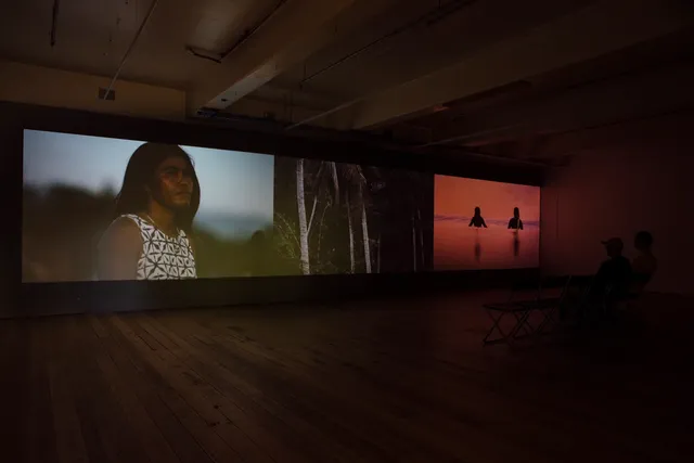 Installation view of a three channel video in the West Space gallery. The gallery has hardwood floors with two people seated on chairs to the right side of the gallery. The first channel shows a Balinese woman with long, dark hair wearing a black and white printed top and gold chain necklace. The blurred background has muted green and blue tones. The second channel shows a forest with three pale tree trunks. The third depicts two female surfers on the water with a vivid pink and red sky.