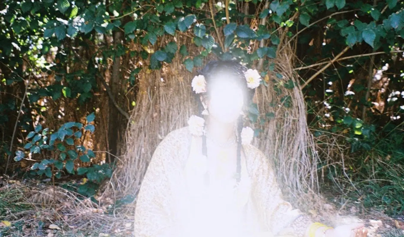 An image of a person in a garment seated on the grass under a tree, they have flowers in their hair and around their neck and are glowing a strong white light.