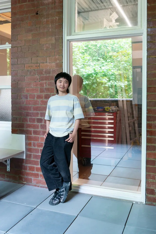 Artist Joy Zhou stands in front of the West Space Window, which they have turned into a see-through mirror, reflecting both outside the Window and making what is inside visible - such as a tool trolley and a ladder.
