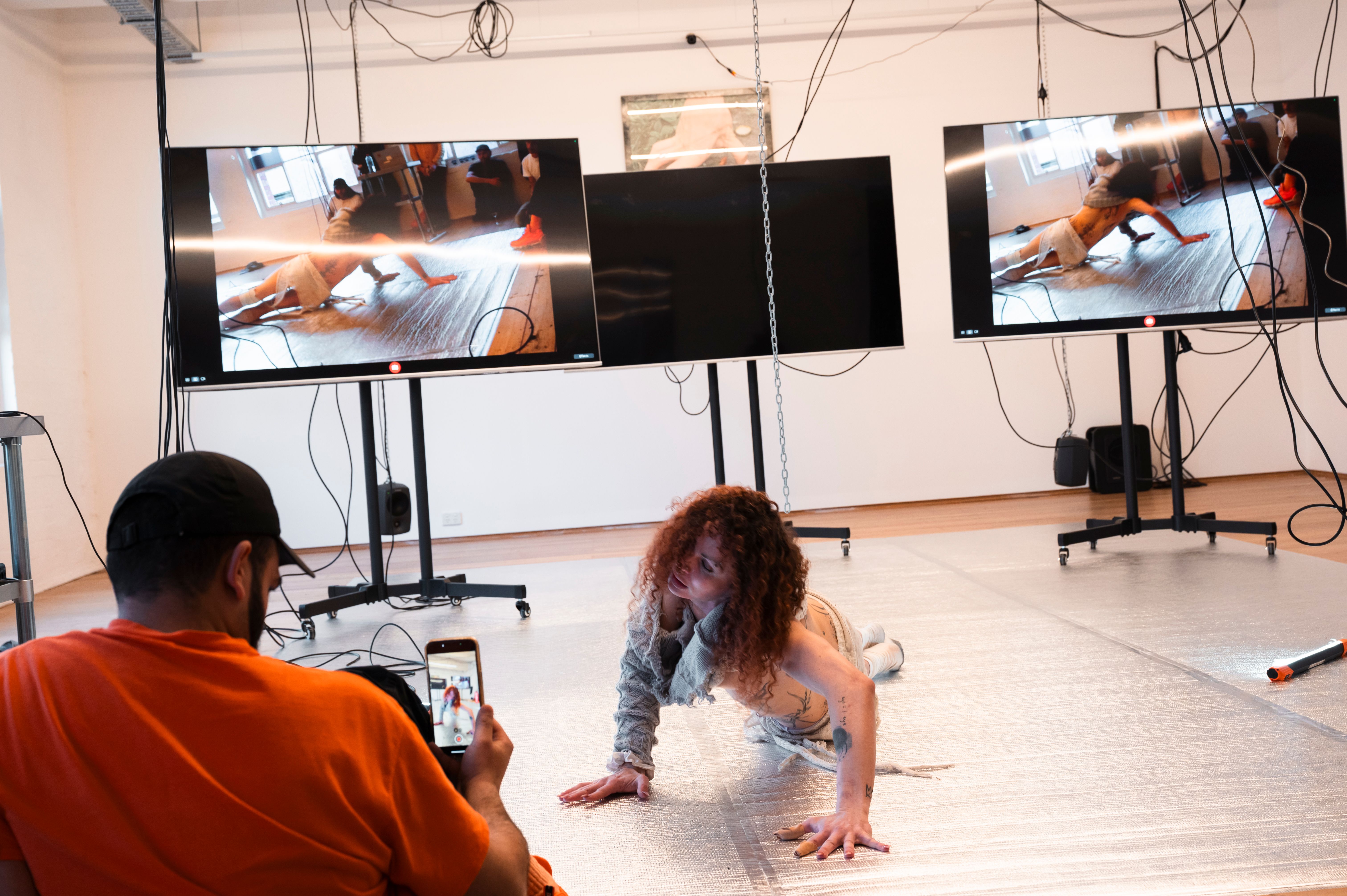 A woman crawls on the silver flooring of the gallery towards audience members recording her on their phones. She is in front of 3 large screens with images of her performing playing on them