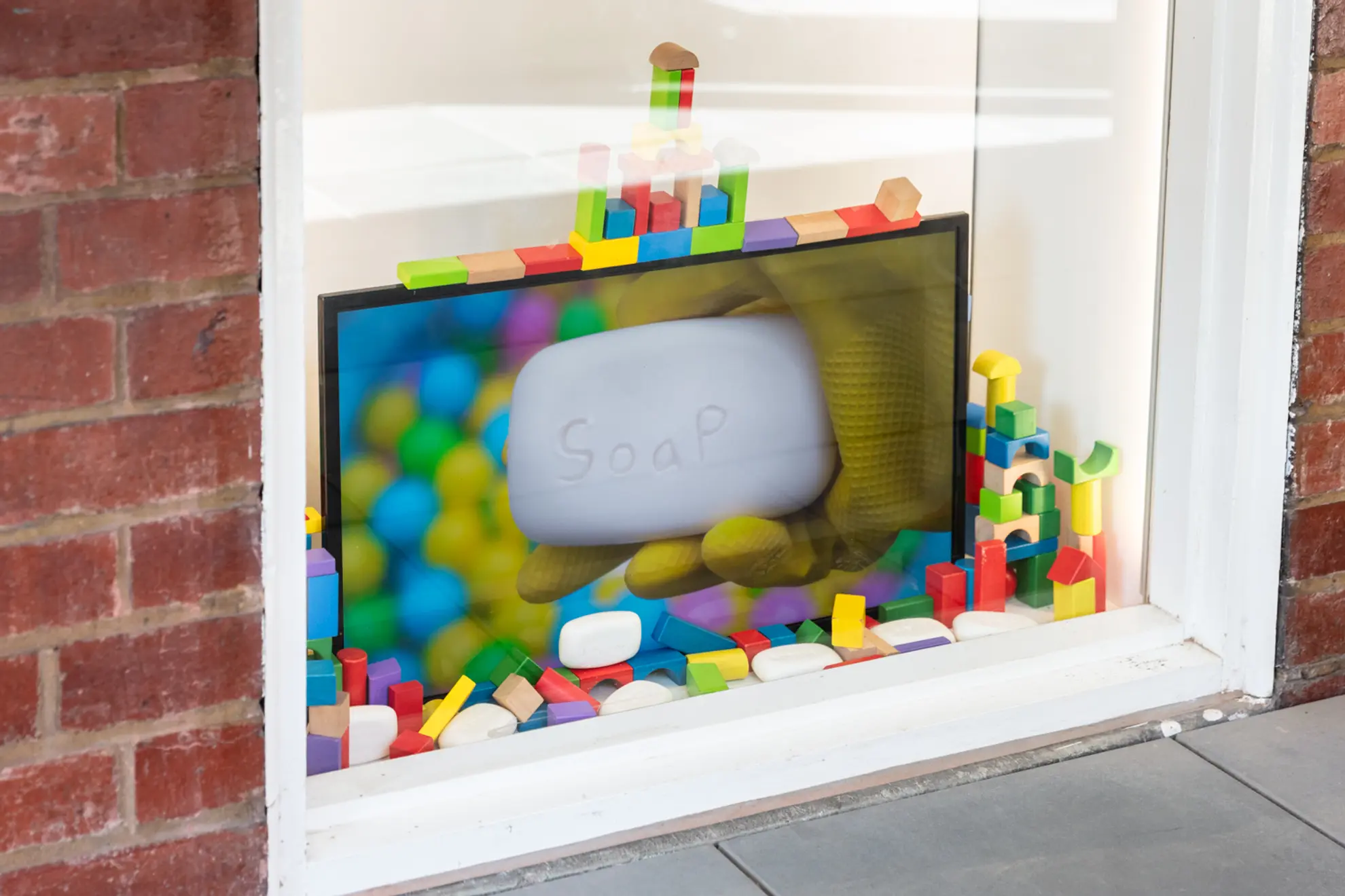 A white window displaying a tv screen with an image of a yellow gloved hand holding a bar of white soap with the word soap engraved onto it. The tv screen is surrounded by other bars of soap and multicoloured wooden brick toys.