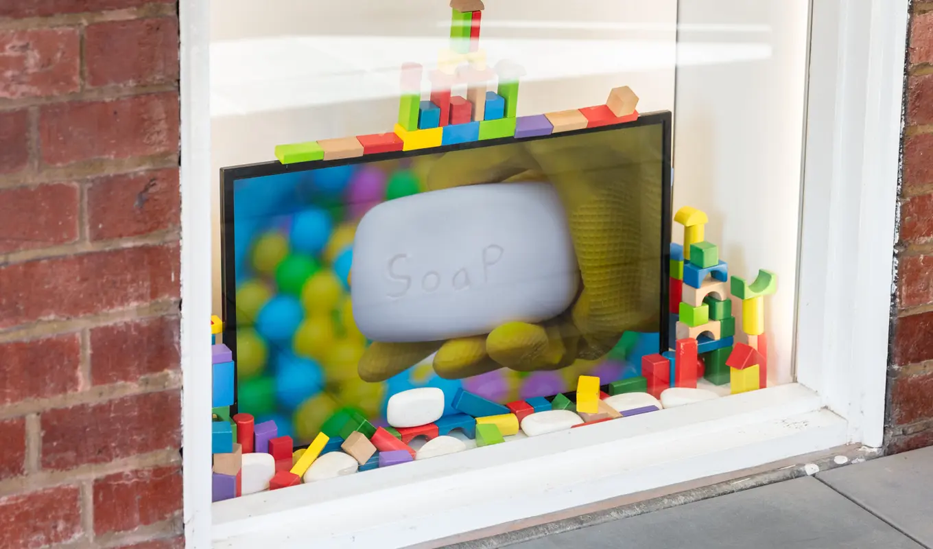 A white window displaying a tv screen with an image of a yellow gloved hand holding a bar of white soap with the word soap engraved onto it. The tv screen is surrounded by other bars of soap and multicoloured wooden brick toys.