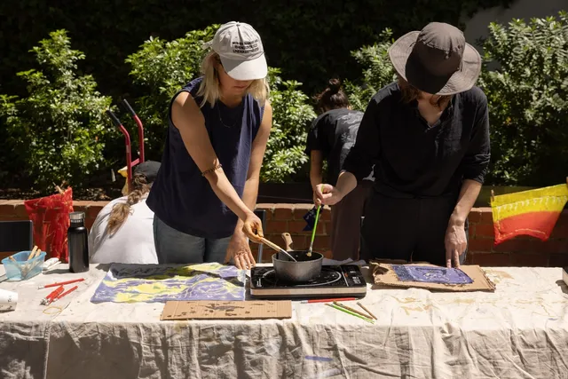 Two figures stand up at a table dipping a brush each into a pot, ready to apply to a surface on the table. They are both wearing hats outdoors, and the table is covered in cloth.