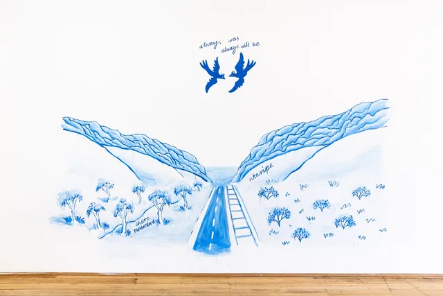 A mural painted in blue paint on a white wall. The image depicts a location known as "the gap" in Alice Springs, Northern Territory, Australia. Two rocky mountain peaks coverge at a low point where a road and train track pass through the middle. There is text that says "there mparntwe". Above there are two birds in flight with text above them that says "always was, always will be"