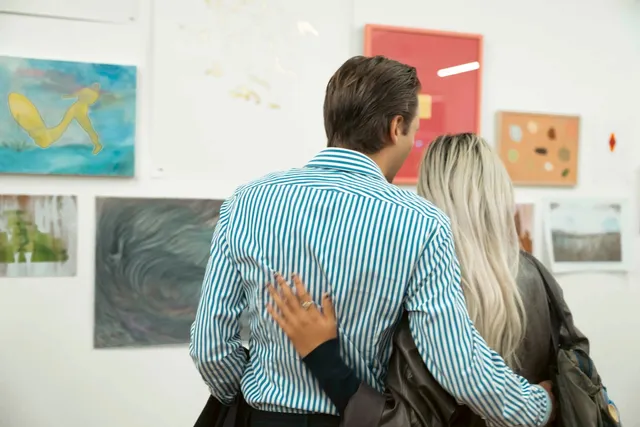 two figures embrace with arms around each other. They are standing in front of an assortment of artworks installed for the West Space fundraiser 2024