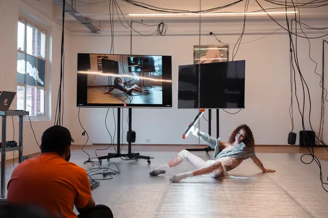 Andrea sits on the floor in front of the audience with her legs spread holding a big light shining between her legs