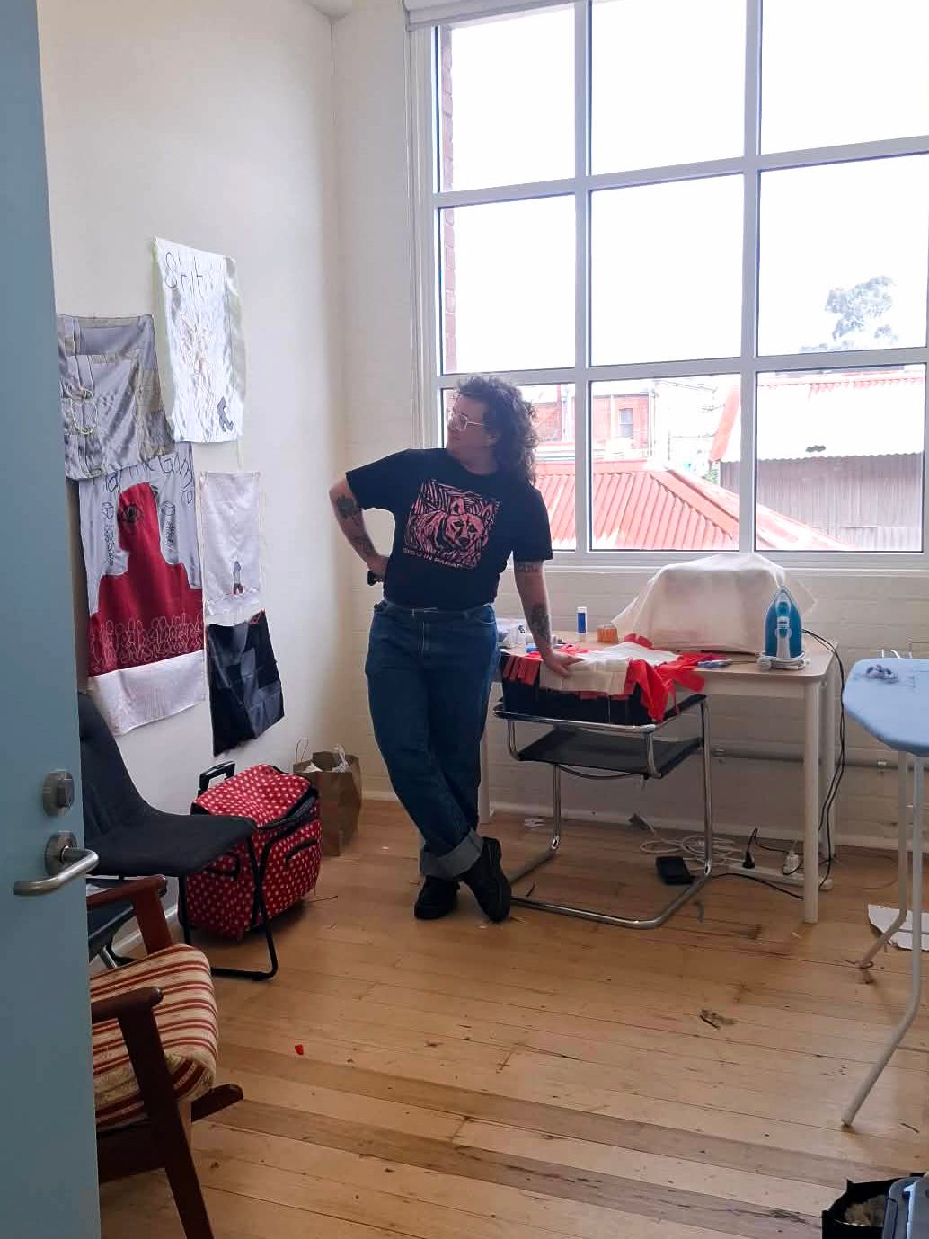 Edwin Devril standing with their legs crossed and a hand on a chair whilst looking to the side towards a wall covered in various textile works.
