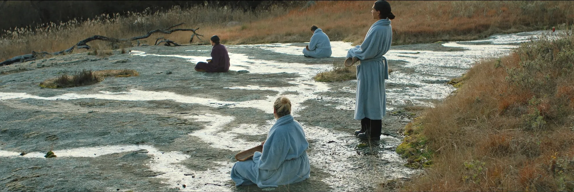 In a large, marshy plain of low level grasses and shrubs we see two people in robes in the distance seated on the floor. There are two more people in the foreground, one is seated and one stands.