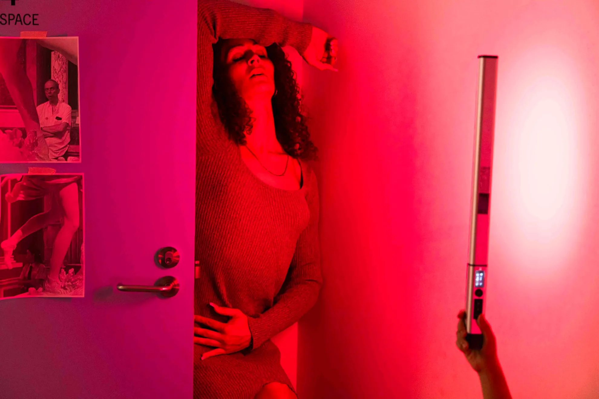 A woman stands behind a door against a wall during a performance. Someone is holding a bright pink light that is shining on her and lighting the room.