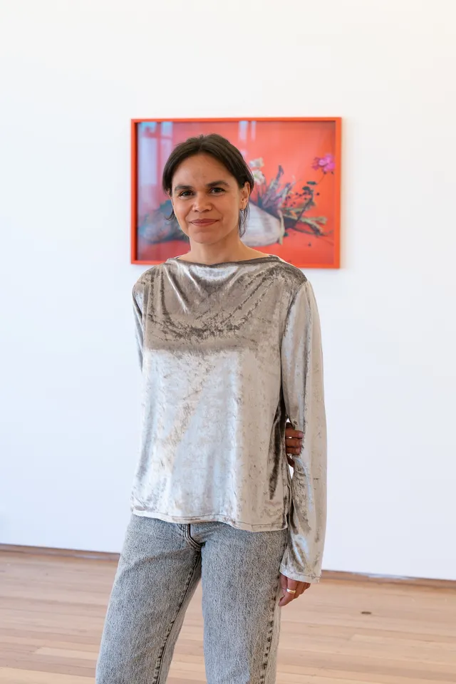 A portrait of Katie West in a suede silver long sleeve top and light wash jeans in front of a framed photography piece. The photograph shows a shell and flowers on a vibrant background.