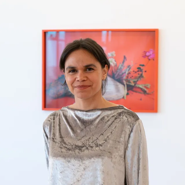A portrait of Katie West in a suede silver long sleeve top and light wash jeans in front of a framed photography piece. The photograph shows a shell and flowers on a vibrant background.