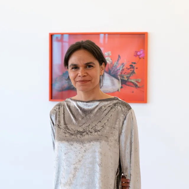 A portrait of Katie West in a suede silver long sleeve top and light wash jeans in front of a framed photography piece. The photograph shows a shell and flowers on a vibrant background.