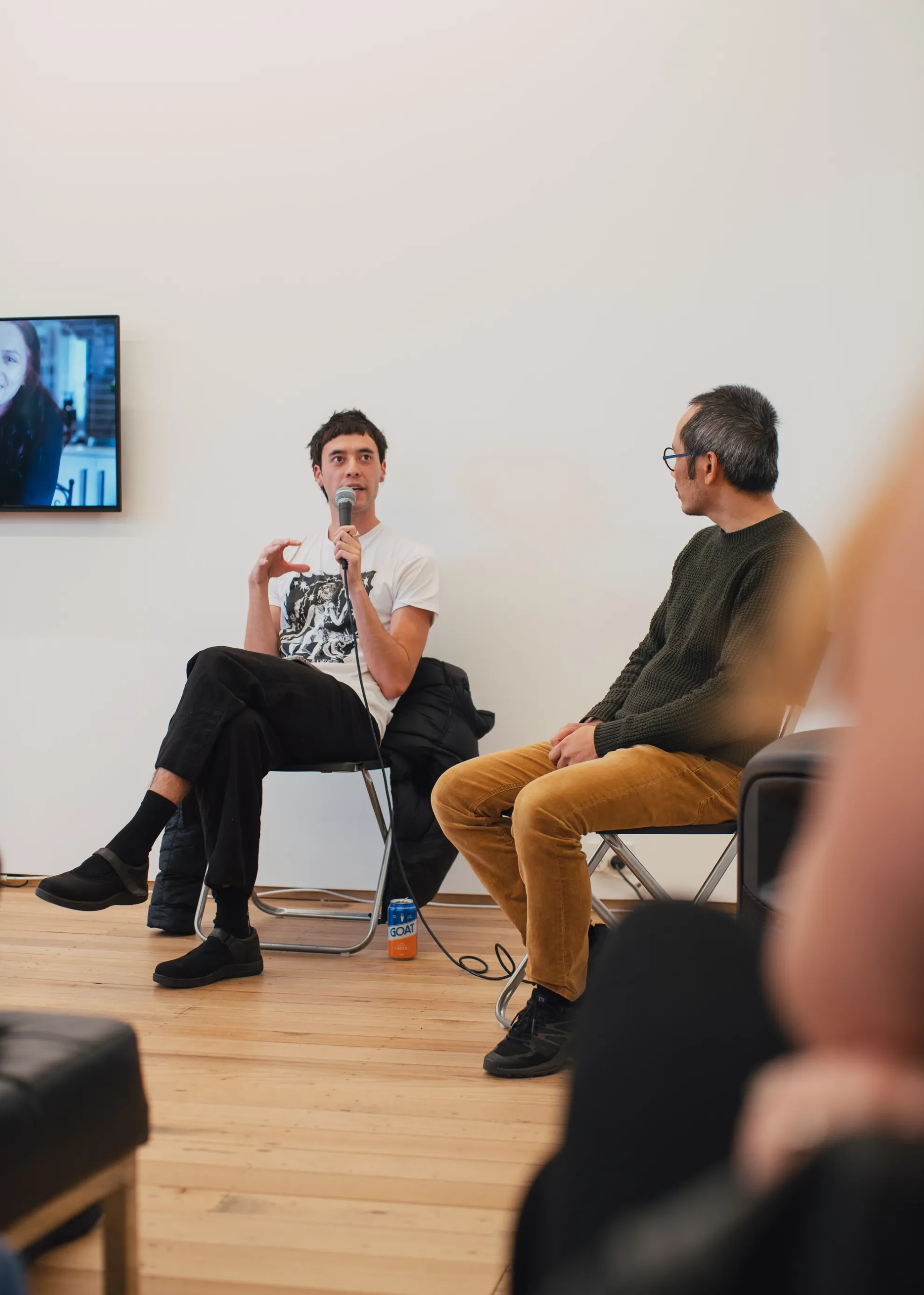 Two individuals sit on fold out chairs. One is sitting crossed-legged speaking into a microphone with a hand out gesturing while speaking. A TV is seen to the far left of him. The other individual sits on the right, intensely listening, with a speaker directly next him. We see the blurred figure of a person in the peripheral listening.