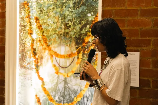 Person speaking with microphone in front of brick wall, window display and small text. Large bright window display with photo prints of trees, beads and orange and yellow flowers hung across the frame.