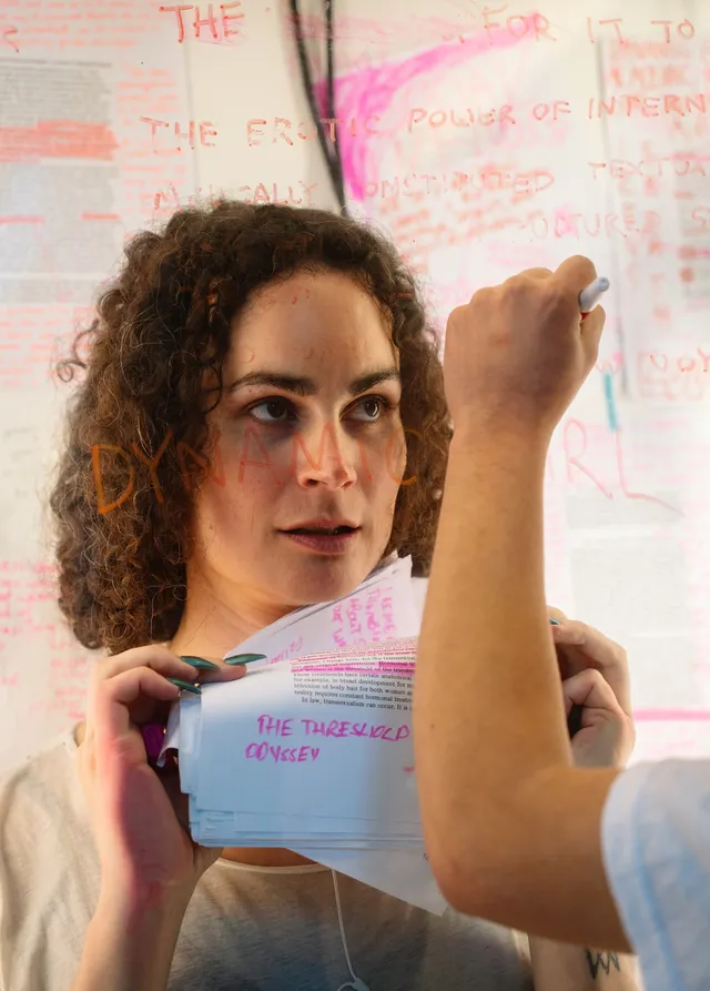 A figure gazing upward behind a window stares at an orange marker as an anonymous person's hand writes the word 'Dynamic' on the window in front of their face. The person behind the window is holding some paper with chunks of printed text and hand written notes in pink highlighter.