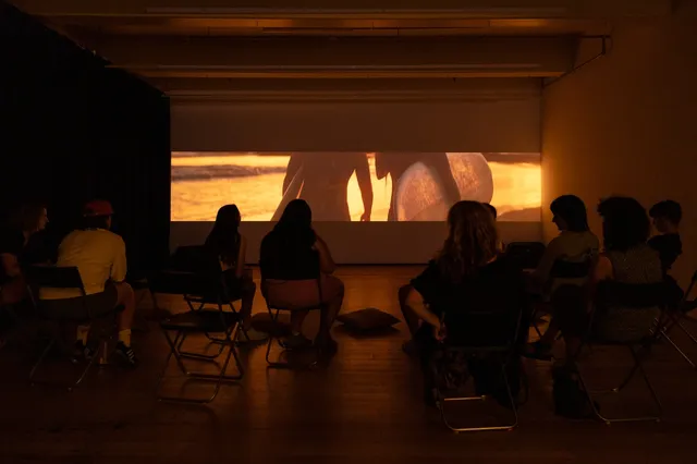 A projection artwork, long and beaming in yellow and orange. There is a crowd of people on chairs looking forward toward the projection.