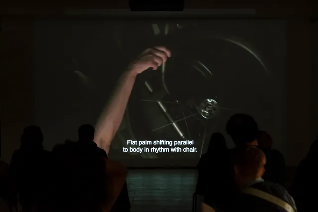 A dark room hosts a group of seated individuals in front of the projector screen. Projector reads 'Flat palm shifting parallel to body in rhythm with chair.' accompanied by a hand turning a wheel.