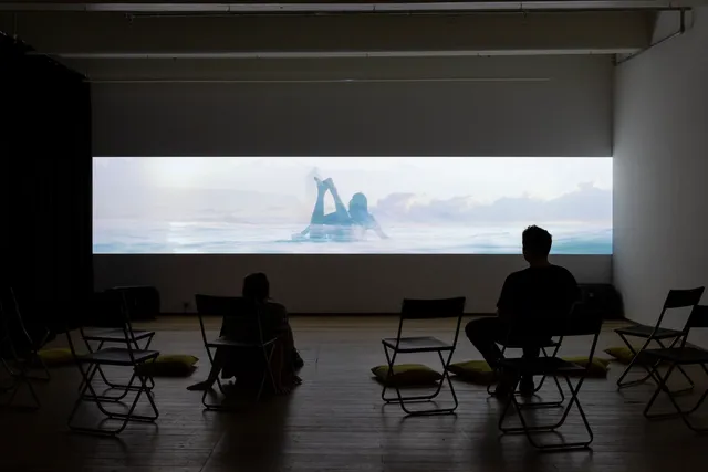 Among some empty chairs and cushions, two people are watching video install.