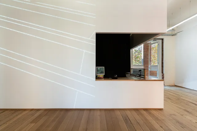 Installation view of "Big Pharmakon". On the right of the image is a countertop with a small analog television on a pile of books and wood and glass structure containing a camera. To the left of the counter a projection is partially visible showing a series of diagonal parallel lines.