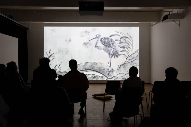 People sitting in a dark room watching a film projected on the white wall. The showing scene is a black and white ink painting.