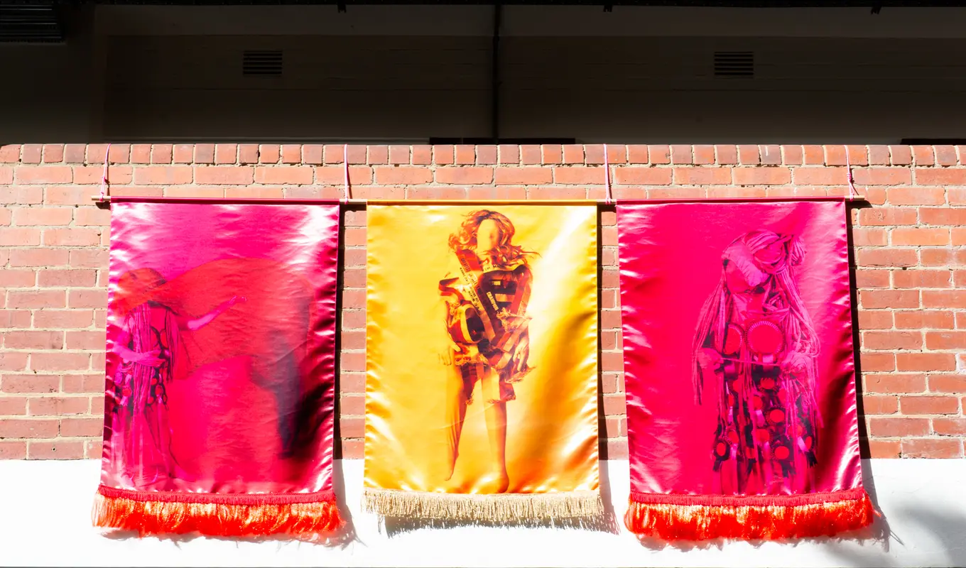 Three draped pieces of fabric over a red brick wall. The two on either side are hot pink and the one in the centre is yellow, all three have various images printed on them.