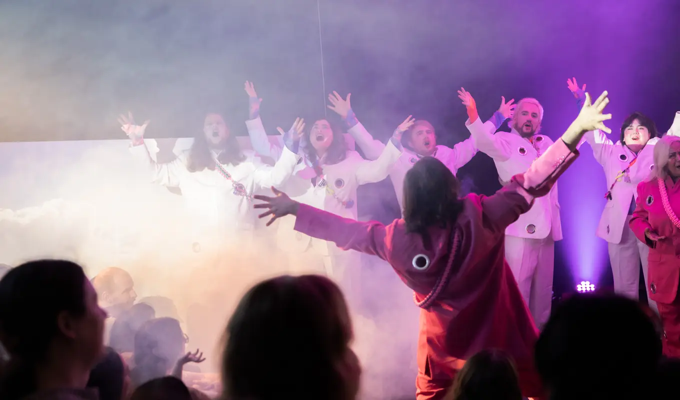 A group of people in white and pink stand amid a smoke machine with jazz hands and chests puffed outward in chorus.
