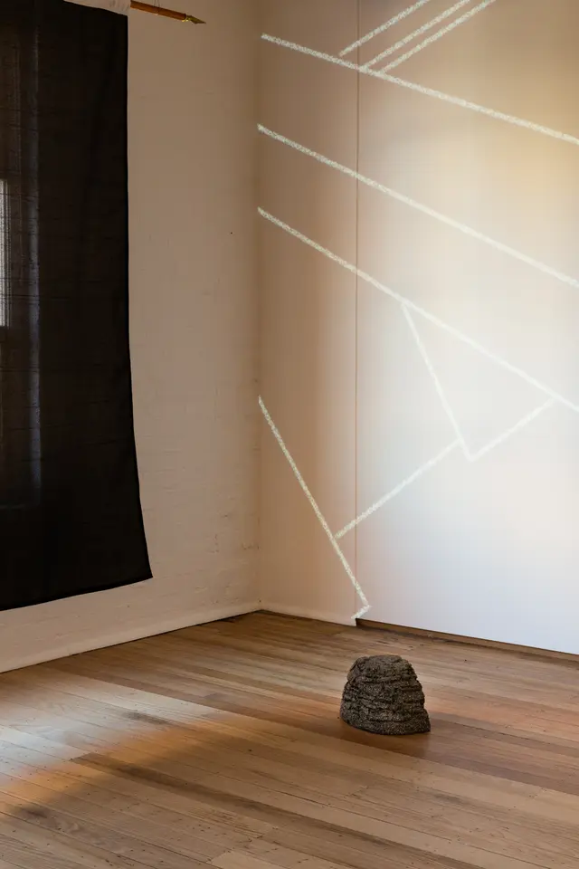 Installation view of "Big Pharmakon". On the left of the image a black semi-transparent sheet is partially visible and suspended from the ceiling. Against the back wall a projection of lines is visible with a small plastic rock sitting in front of it.