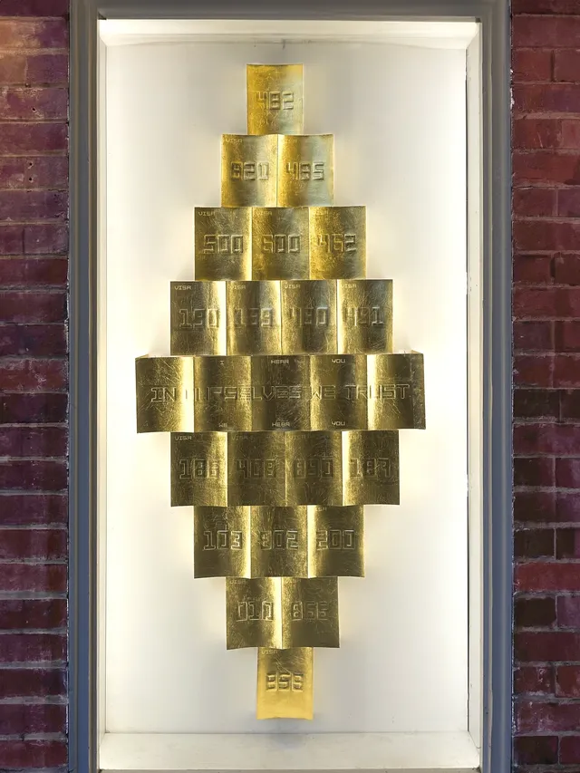 A close up installation view of the west space window. There are a collection of gold rectangular objects that are arranged in a diamond formation. These shapes are emobossed with 3 digit numbers representing the immigration visa codes in Australia