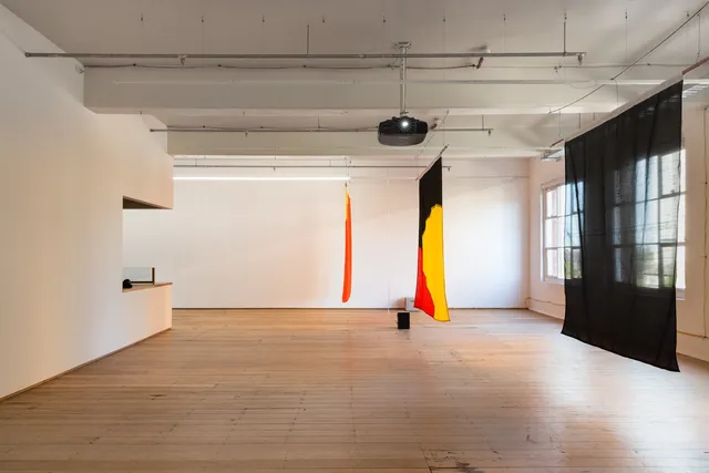 Installation view of "Big Pharmakon". The image is a side on view of 3 semi transparent sheets suspended in the air in the West Space Gallery. The closest sheet is completely black with the back two showing red, yellow and black shapes that are partially visible. A speaker is visible between the sheets on the floor and a projector faces the viewer hanging from the roof in the centre of the image.