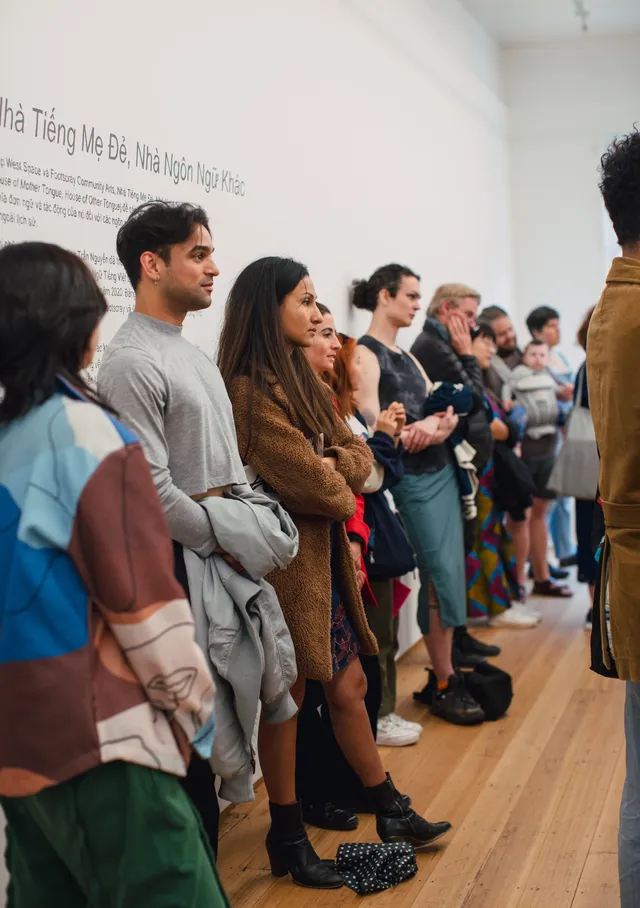 A long line of people leaning agains a white wall from a side on perspective. Behind one of the more foregrounding figures there is a large body of text on the wall. Everyone is looking toward something ahead of the wall.
