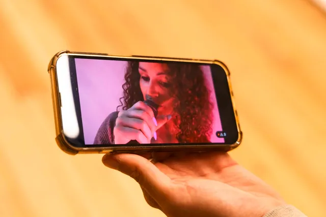 A hand holds an iphone showing a livestream of a figure holding a microphone in pink light. She has long white nails and long curly dark hair.