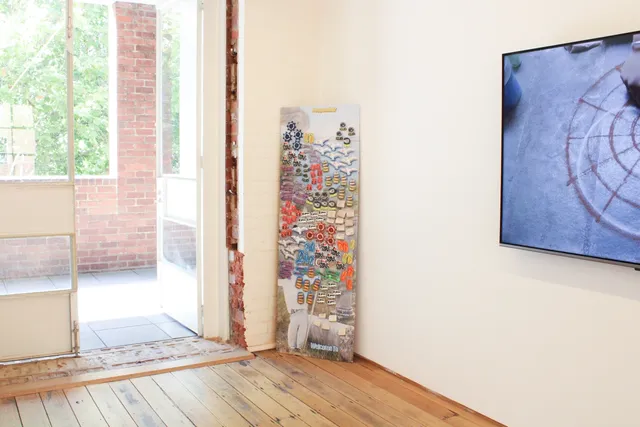 a narrow piece of wall in the corner of the gallery facing outward. Attached are an array of magnet-like forms, in multiples and different beach-related shapes. This includes seagulls, thongs, ship wheels, and magnets that say 'I love philosophy'. On the surface of the wall-like form there is a figure with open arms in the bottom left corner and text reading 'Welcome to'.