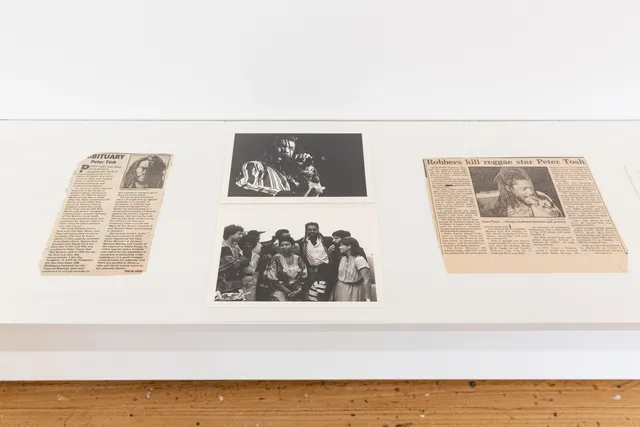 The photo shows 2 newspaper clippings and 2 photos sitting on a white shelf suspended off of the wall. The news paper clippings report on the death of Peter Tosh and the 2 black and white photos are of Tosh performing and Tosh surrounded by a group of people.