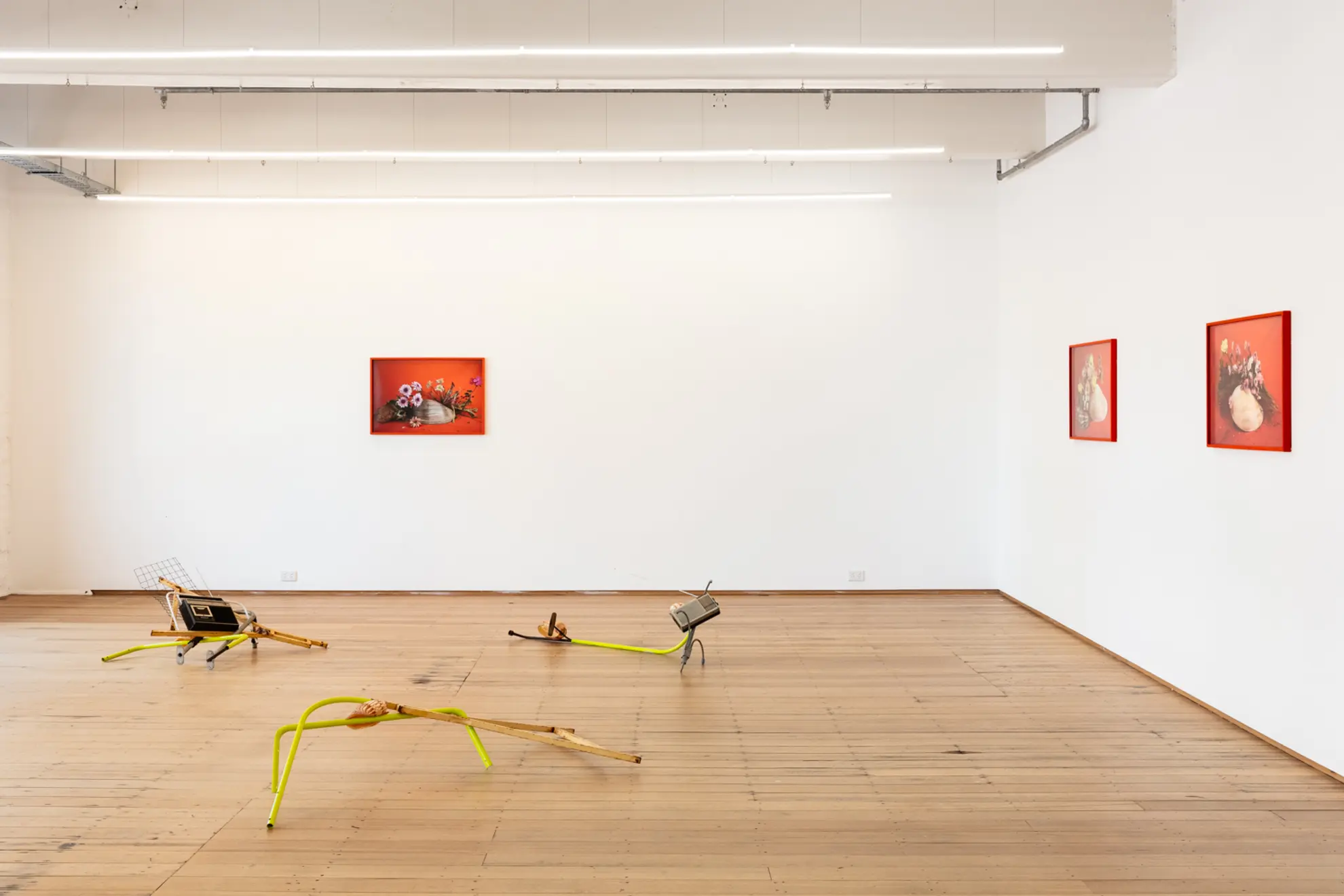 Three of the sculptural installation pieces sit on the gallery floor comprised of metal poles and radios, in the background three framed photography pieces hang from the walls of flowers resting on shells.