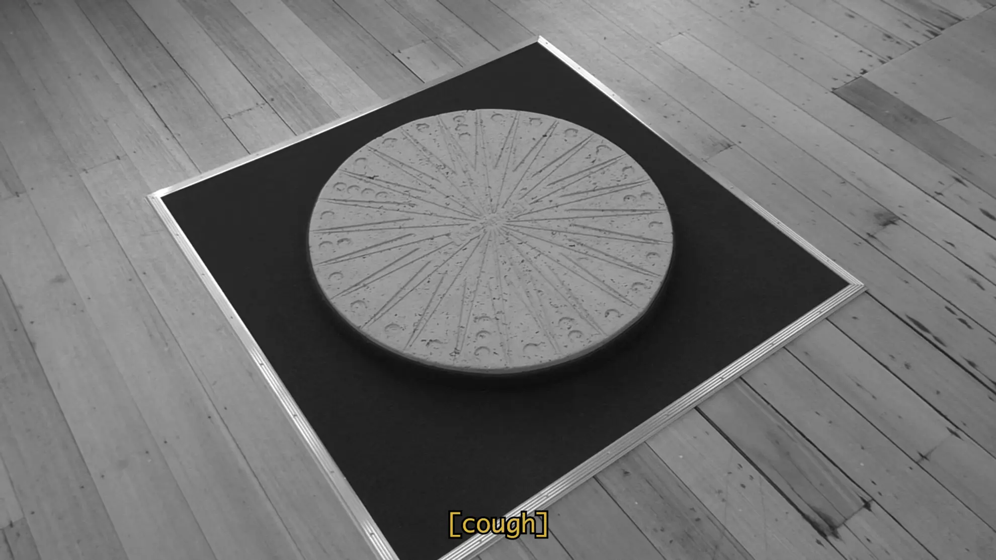 a black and white film still of a large concrete circular disc that resembles a man hole cover sits of a square of carpet on top of floorboards.