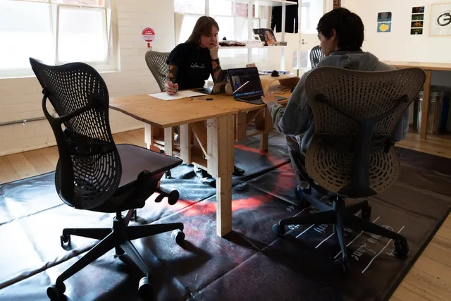 2 gallery staff sit around a long wooden table that sits on top of a large black and red floor based vinyl artwork by Diego Ramirez