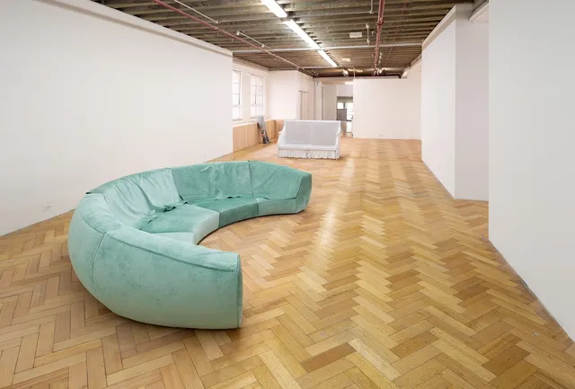 Installation view of 'Rose Moon', an exhibition by Fiona Abicare. There are three sculptures in the space. A moon shaped longe covered in teal fabric is the focal point of the image. A second two seater couch upholstered in white denim sits behind the first sculpture. A sculpture of a pair of legs in denim jeans and boots leans against the gallery wall in the background of the image.