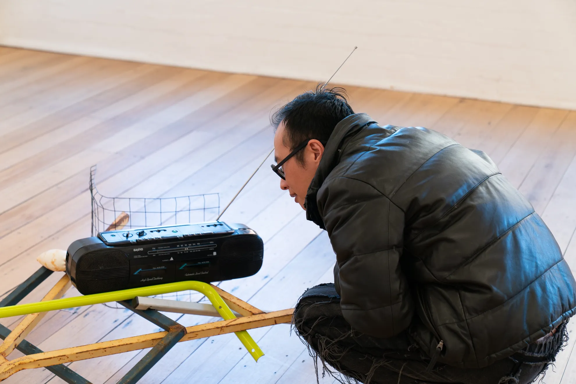 A person in a shiny puffer jacket with glasses leans closely to the radio component of the sculptural installation work. The radio rests on multiple metal poles and is supported by a wire frame.