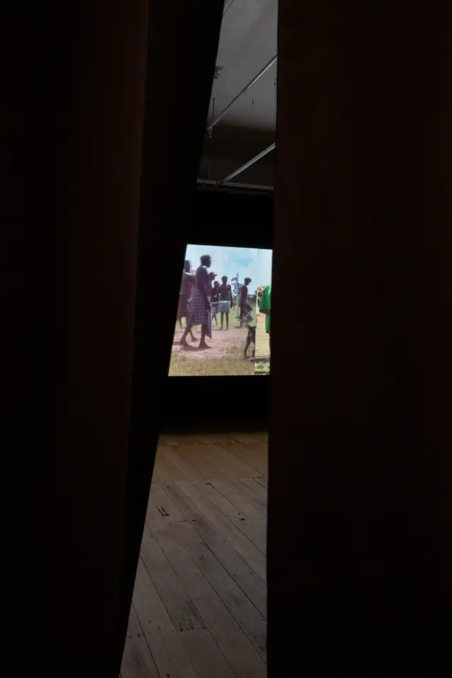 Two black curtains have parted slightly to show piece of a film still projected on the gallery wall. The image shows a small gathering of people standing and walking outside on grass and dirt.