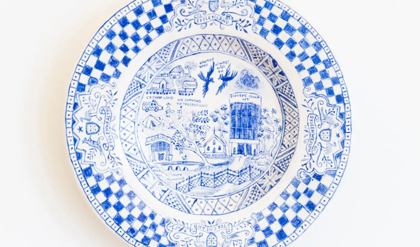A hand painted ceramic plate in blue on white. The outer rim of the plate is a thick checkered pattern. The inner of the plate is of intricate, detailed subject matter that faintly depicts segments of a landscape.
