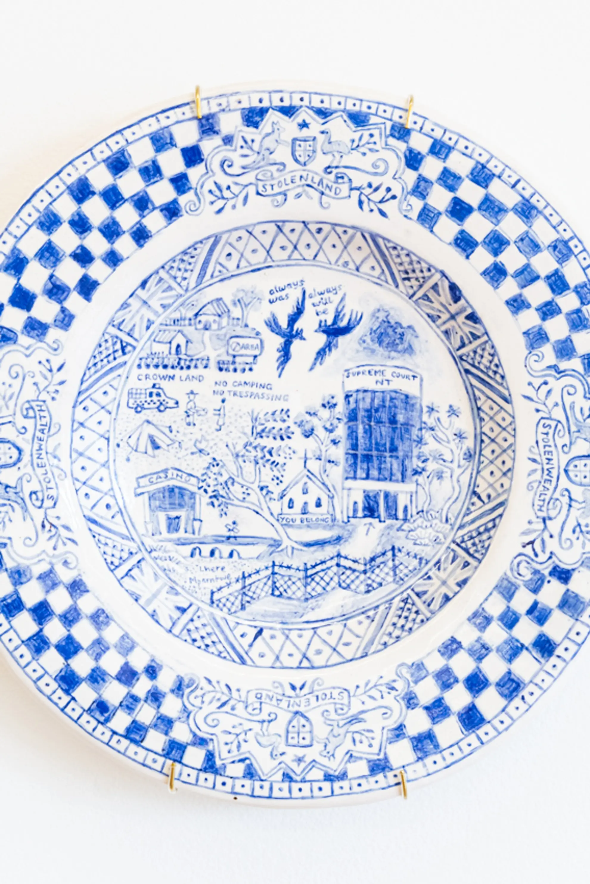A hand painted ceramic plate in blue on white. The outer rim of the plate is a thick checkered pattern. The inner of the plate is of intricate, detailed subject matter that faintly depicts segments of a landscape.