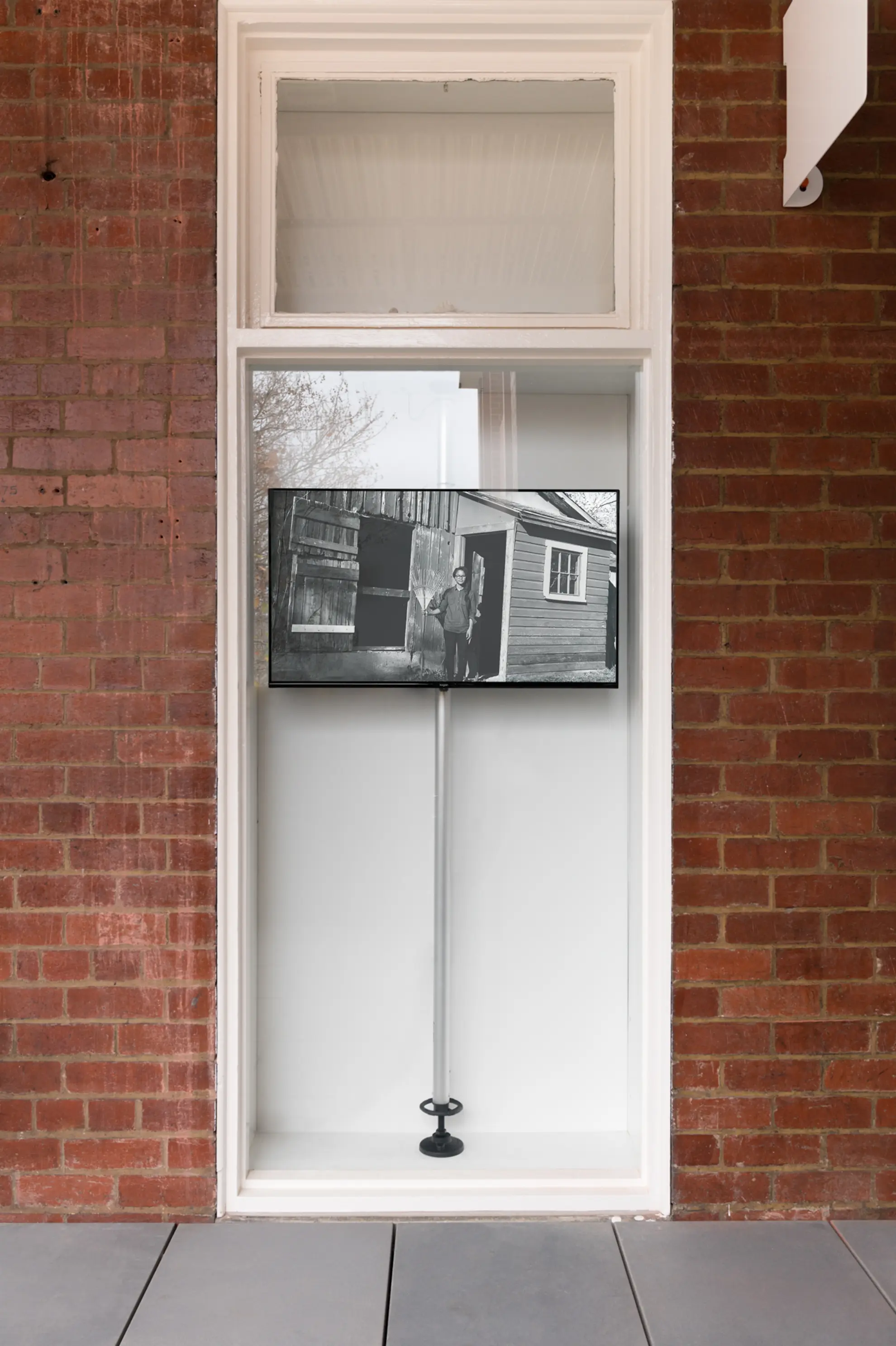 Straight on view of the West Space window. In the top section of the large window pane, is a rectangular screen showing a black and white image of a man standing outside a wooden house. The screen is sitting on a silver pole with a black base.