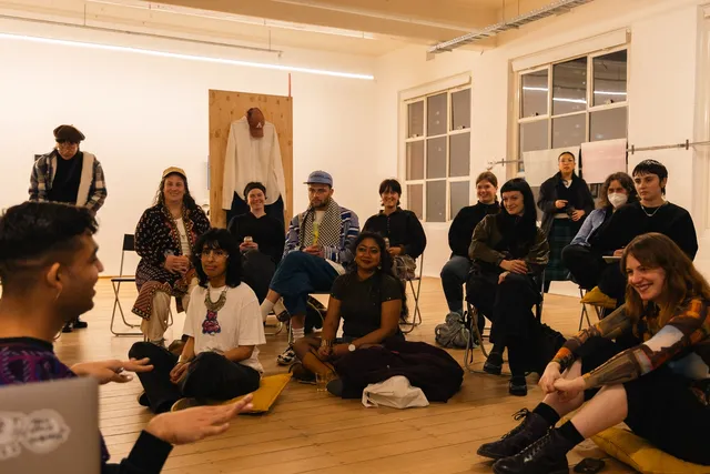 A group of people sit in the floor space of a gallery, some seated on folding chairs, others seated on the floor on top of yellow cushions. They're listening intensely and smiling at one individual who is speaking with his hands out. There are two large windows behind the crowd, as well as an installed wood panel that features a shirt, pants and a hat.