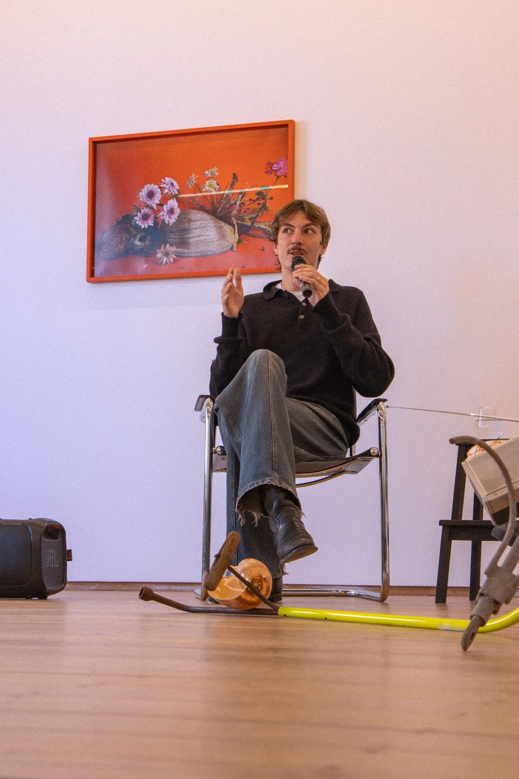 Michael is seated mid speech holding the microphone. He wears a black jumper and dark jeans, behind him we see a framed photographic piece by Katie West of flowers resting on a shell.