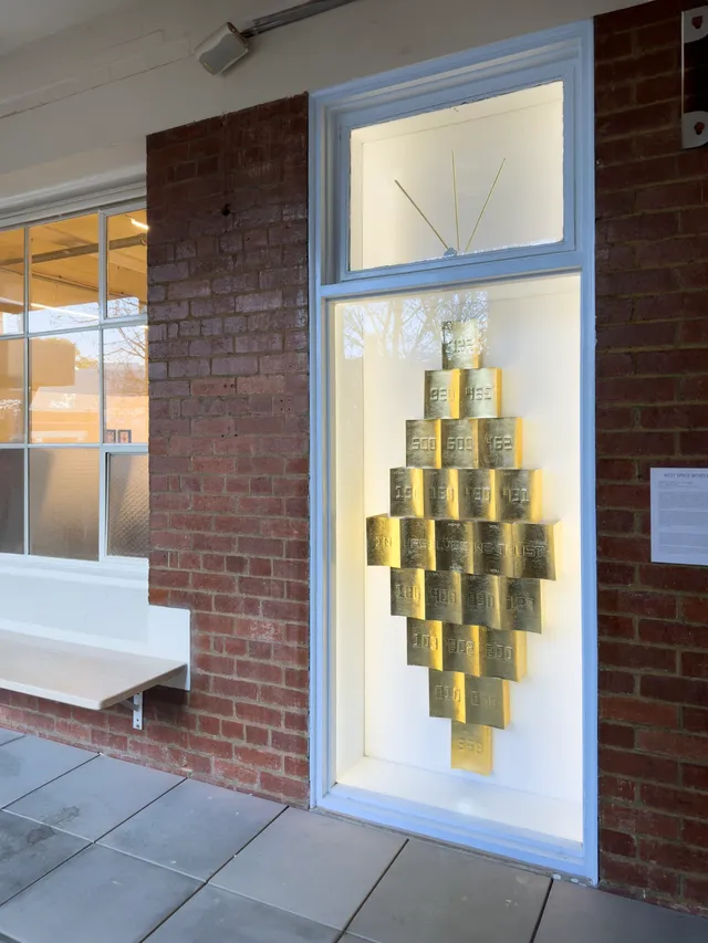 A white walled window space is filled with gold rectangular objects. The window is surrounded by bricks and other windows.