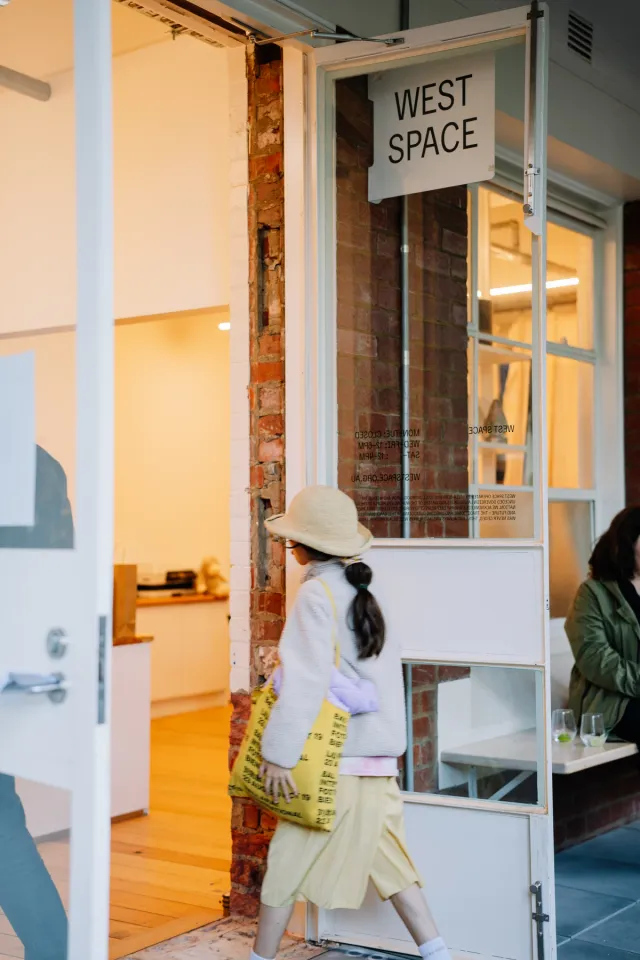 A young figure with a yellow tote bag and big floppy sun hat is entering through the big open doors of West Space gallery below a sign that says "West Space"