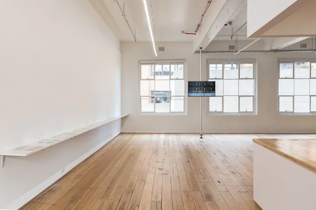 The image is of a screen suspended at eye level in the West Space Gallery showing Guled Abdulwasi's "The Block" (2019). On the left hand side is a shelf suspended off of the wall displaying a series of photographs.